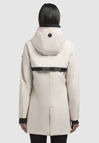 Veste beige résistante à l'eau avec capuche, dotée d'une sangle de maintien noire à l'arrière et de poignets noirs. Texture lisse et coupe ajustée.