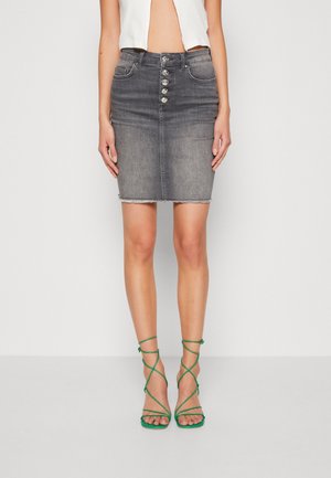Kvinde iført en falmet sort denimnederdel med knapper foran og grønne sandaler med hæl og stropper, der står mod en ensfarvet baggrund.