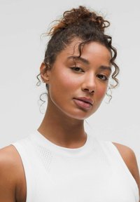 Biały sportowy top na ramiączkach z wysokim dekoltem i perforowanymi detalami. Gładka tekstura, z logo marki z przodu. Kręcone włosy ułożone w górę.