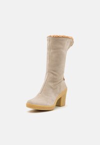 Bota de ante beige hasta la rodilla con un cuello texturizado y suave, tacón en bloque y suela de goma contrastante. Presenta un detalle de costura en los laterales.