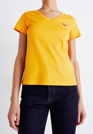 Keltainen lyhythihainen T-paita, jossa V-pääntie, valmistettu puuvillasta. Vasemmassa rinnassa pieni punainen logo, yhdistetty tummiin farkkuihin.