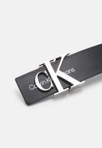 Cintura in pelle nera con una fibbia in argento con logo Calvin Klein. Testo "Calvin Klein Jeans" stampato sulla cinghia. Texture liscia, design minimalista.