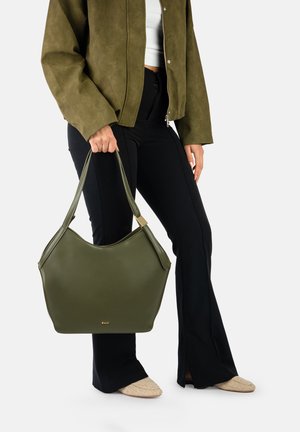Personne tenant un grand sac à main vert olive, portant une veste vert olive, un haut blanc, un pantalon noir et des chaussures beiges, debout devant un fond uni.
