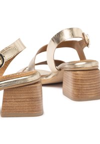 Sandali metallici dorati con tacco a blocco, caratterizzati da una base in legno testurizzato e un design a doppia cinturino con chiusura a fibbia regolabile.