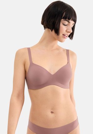 Donna con i capelli corti e scuri che indossa un reggiseno malva senza cuciture e biancheria intima abbinata, che guarda in basso verso sinistra su uno sfondo bianco.