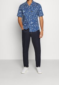 En person står självsäkert, iklädd en blåmönstrad kortärmad skjorta och mörka jeans, tillsammans med vita sneakers, framför en neutral bakgrund.