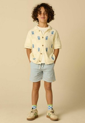 Enfant aux cheveux bouclés, les mains dans les poches, portant un cardigan crème avec des homards bleus, un short rayé, des chaussettes à carreaux et des baskets.