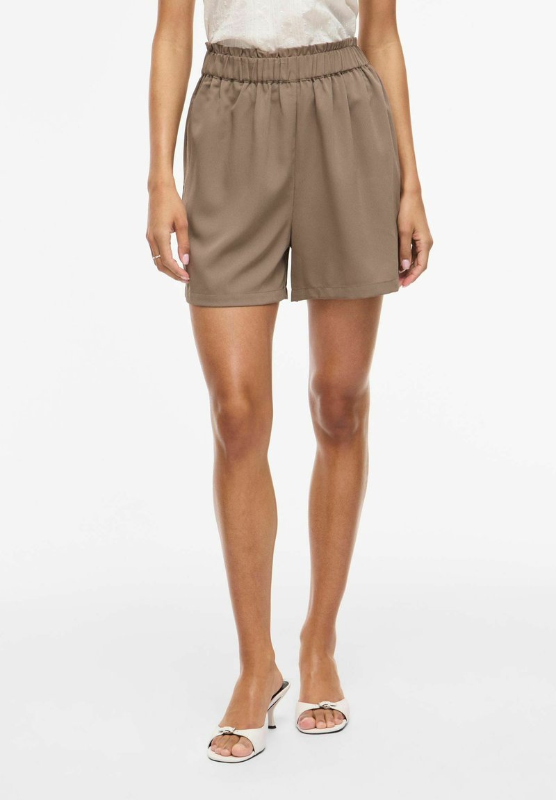 Vrouw die hooggetailleerde bruine elastische shorts en witte open-toe sandalen met hak draagt, staand tegen een effen lichte achtergrond.