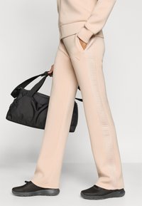 Μπεζ παντελόνι φούτερ με στενή γραμμή, με υφασμάτινη πλαϊνή λωρίδα με λογότυπο, σε συνδυασμό με μια μαύρη τσάντα duffel και μαύρα παπούτσια από δίχτυ.