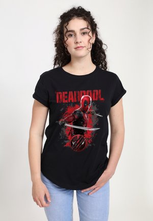 Femme aux cheveux bouclés portant un t-shirt noir Deadpool, tenant un katana et un jean bleu clair sur un fond blanc uni.
