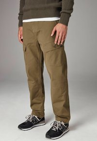 Khaki cargobroek met meerdere zakken, gemaakt van stevige katoenstof, heeft een rechte snit en versterkte knieën. Ggedragen met zwarte sportschoenen.