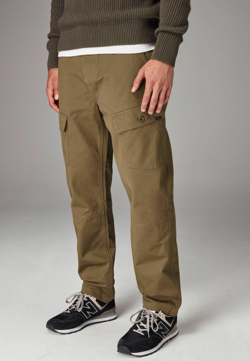 Khaki cargobroek met meerdere zakken, gemaakt van stevige katoenstof, heeft een rechte snit en versterkte knieën. Ggedragen met zwarte sportschoenen.