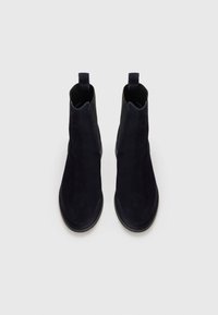 Botas de tobillo de ante en azul marino oscuro con paneles laterales de elástico, punta redondeada y tirador, que presentan una textura suave y un diseño minimalista.