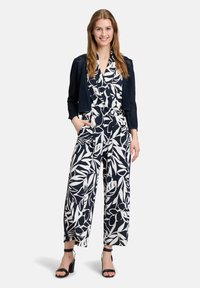 Navy- und weiße Blumen-Jumpsuit mit weitem Bein, Wickel-Front und seitlichen Taschen, kombiniert mit einer navyblauen Cropped-Strickjacke und schwarzen Blockabsatzsandalen.
