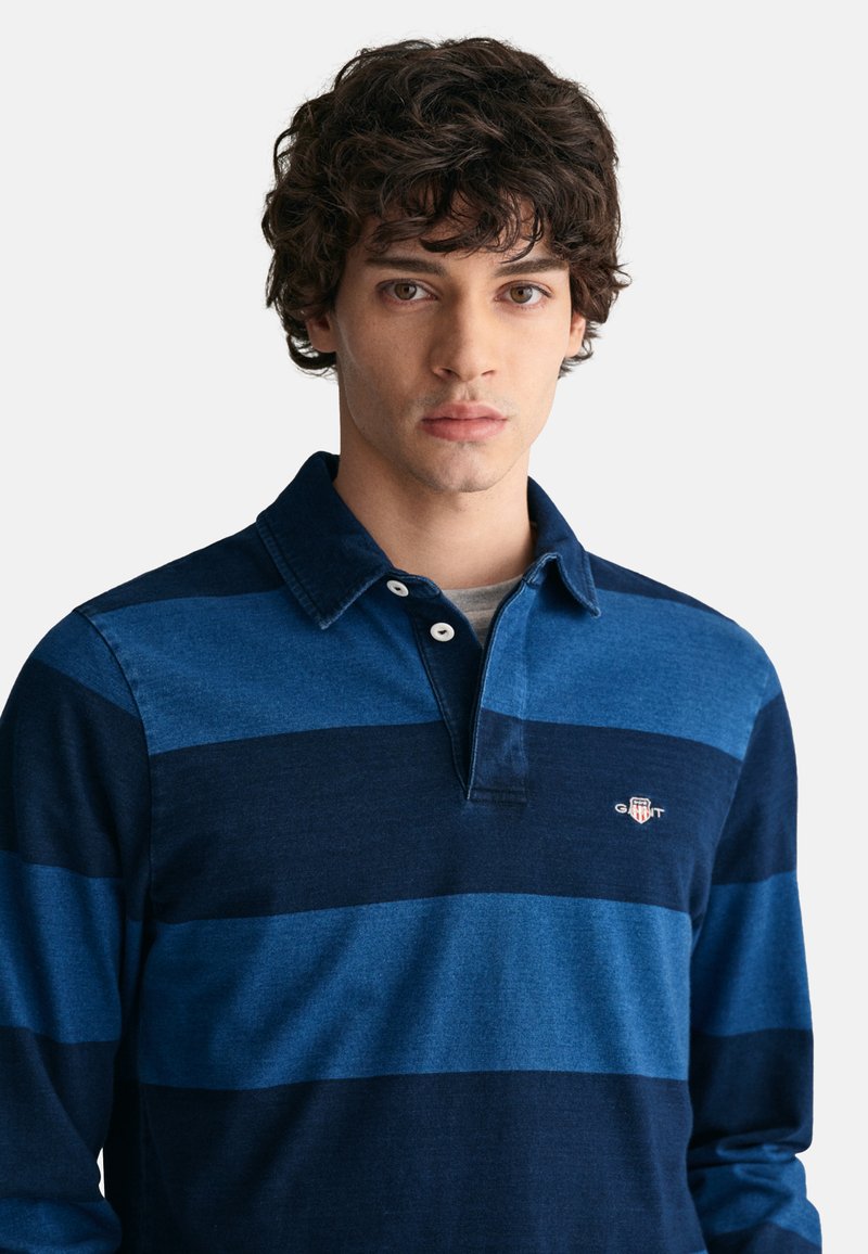 Giovane uomo con capelli ricci scuri che indossa una polo a maniche lunghe a righe blu e blu navy con un piccolo logo sul petto.