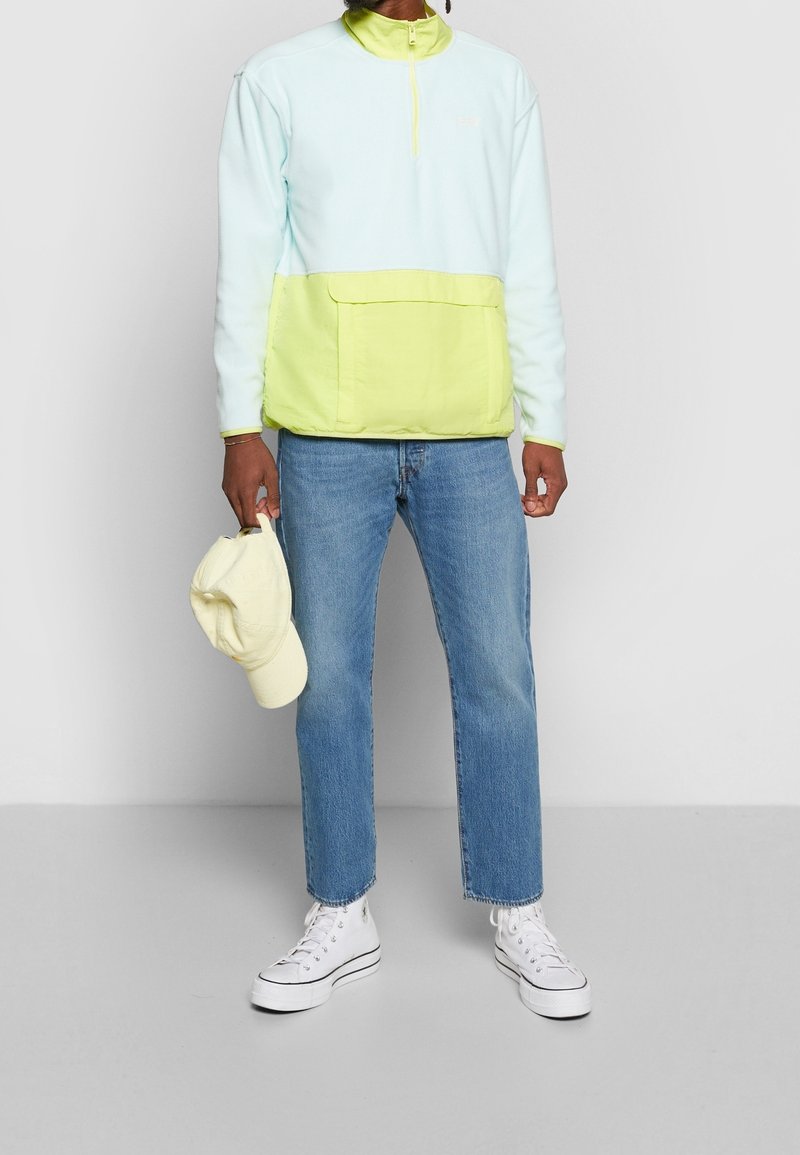 Pullover azzurro chiaro, design a mezza zip, con una tasca giallo neon a contrasto; indossato con jeans blu e sneaker alte bianche; cappellino giallo pallido.