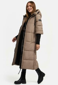 Langer beiger Daunenmantel mit Kapuze, kurzen Ärmeln, Reißverschluss und Seitentaschen. Über dunkler Kleidung mit schwarzen Stiefeletten getragen.