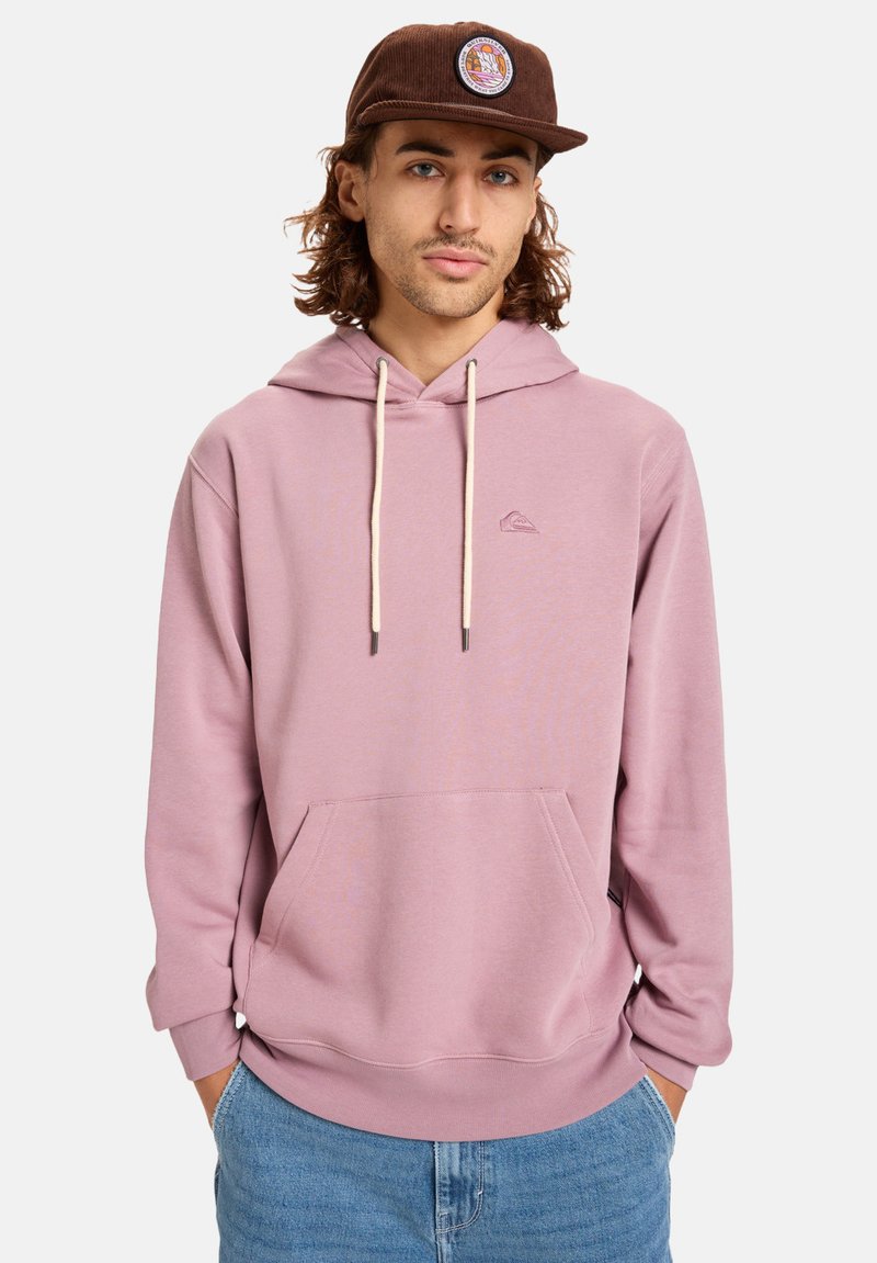 Sudadera rosa claro con un bolsillo frontal, capucha con cordones y un sutil detalle de logo. Combinada con jeans azules y una gorra marrón.