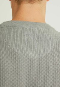 Grå stickad tröja med en texturerad mönster, ribbad halsringning och en subtil broderad detalj på baksidan. Standard passform.