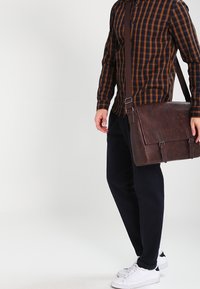 Sac messager en cuir marron avec fermeture à double boucle, bandoulière réglable et finition texturée. Porté avec un pantalon noir et des baskets blanches.