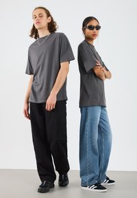 Zwei Personen tragen lässige Kleidung: ein graues T-Shirt, schwarze Cargo-Hosen und blaue weit geschnittene Jeans. Eine Person trägt dunkle Sonnenbrillen.