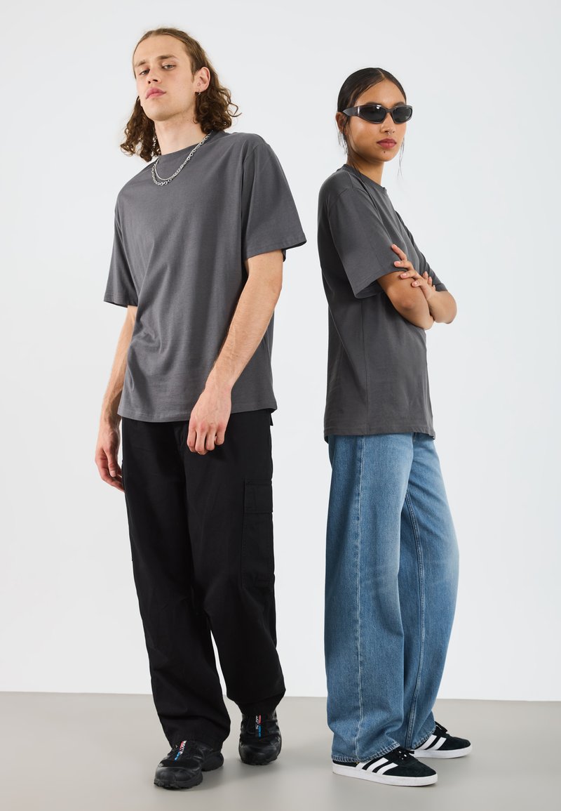 Zwei Personen tragen lässige Kleidung: ein graues T-Shirt, schwarze Cargo-Hosen und blaue weit geschnittene Jeans. Eine Person trägt dunkle Sonnenbrillen.