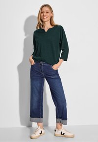 Haut texturé vert foncé avec un col en V, associé à un jean en denim bleu ample, retroussé aux chevilles, et des baskets blanc cassé.