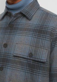 Camicia a quadri in tonalità di blu e grigio, con taschino frontale, bottoni neri e un tessuto testurizzato. Presenta un design classico del colletto.