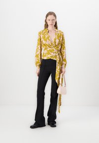 Gelbe floral bedruckte Wickelbluse mit langen Ärmeln, V-Ausschnitt und Bindedetail, kombiniert mit schwarzer Slim-Fit-Hose und einer kleinen pinken Handtasche.