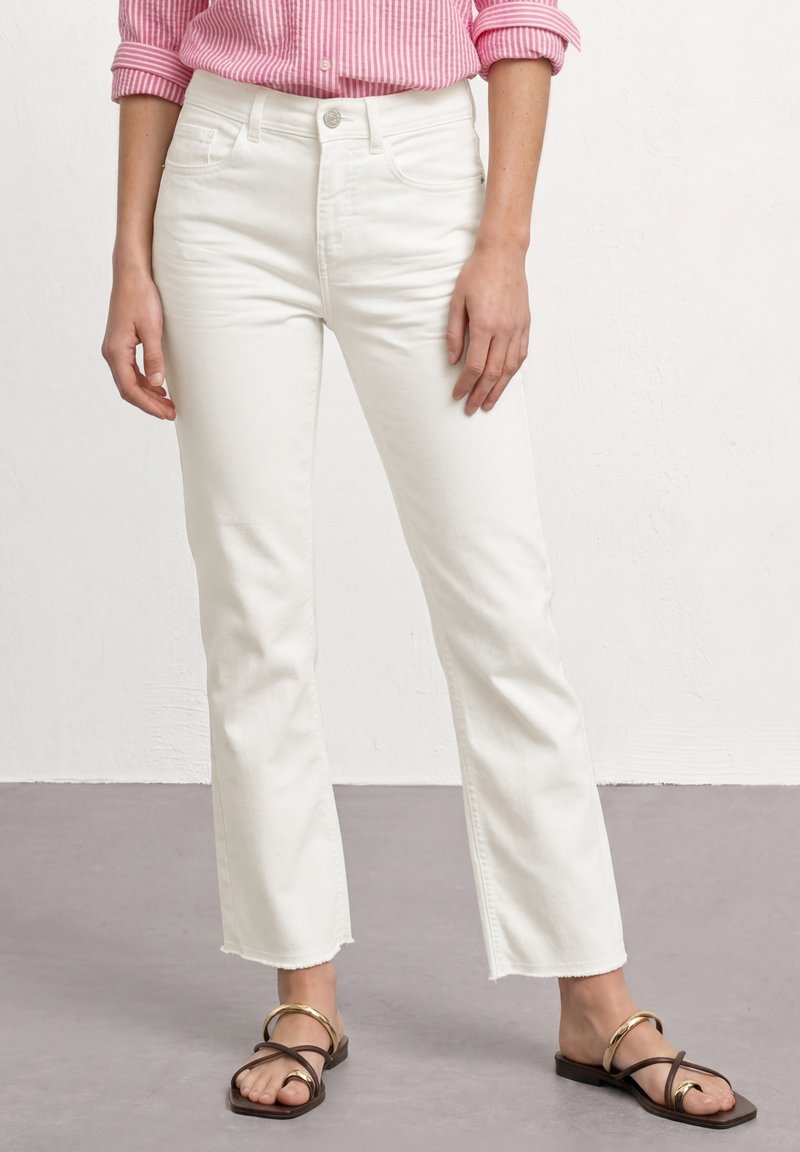 Personne portant un jean blanc court, une chemise à manches longues rayée rose et blanche, et des sandales plates noires avec des brides dorées, debout sur un sol gris.