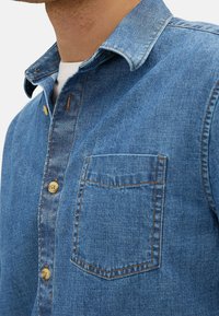 Camicia in denim di blu medio con colletto a punta, chiusura a bottoni e taschino sul petto. Presenta bottoni beige e cuciture a contrasto.