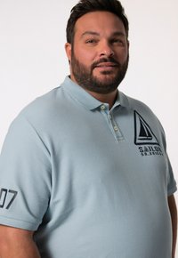 Světle modrá polo košile vyrobená z bavlny, s námořnickou lodí v logu na hrudi a číslem "07" na levém rukávu.