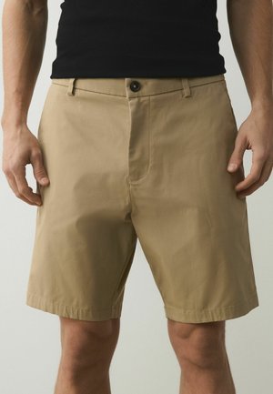 Bărbat purtând pantaloni scurți bej până la genunchi și un top negru fără mâneci, stând cu mâinile relaxate pe lângă corp, în fața unui fundal simplu.