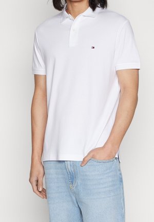 Homme portant un polo à manches courtes blanc avec un petit logo sur la poitrine et un jean bleu clair, main dans la poche.
