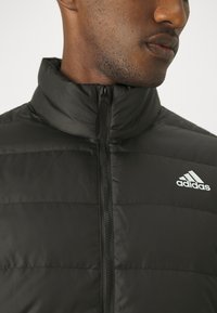 Crna podstavljena jakna s visokim ovratnikom, zatvaračem sprijeda i srebrnim Adidas logotipom na lijevoj strani prsiju. Glatka tekstura i moderni dizajn.