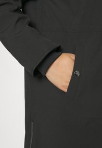 Veste noire avec une texture douce, dotée d'une poche zippée cachée et d'un rabat boutonné. Les poignets présentent un détail en tricot contrastant.
