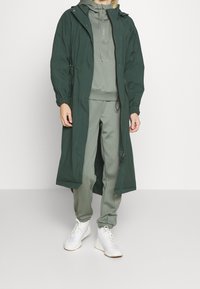 Dlouhý zelený nepromokavý kabát s kapucí na stažení, navrstvený přes odpovídající zelený sportovní oblek, doplněný bílými sportovními botami.