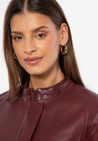 Bordeaux leren jas met een hoge kraag en ritssluiting. Heeft een gladde textuur en minimale stikseldetails. Gouden ringoorbellen zijn zichtbaar.