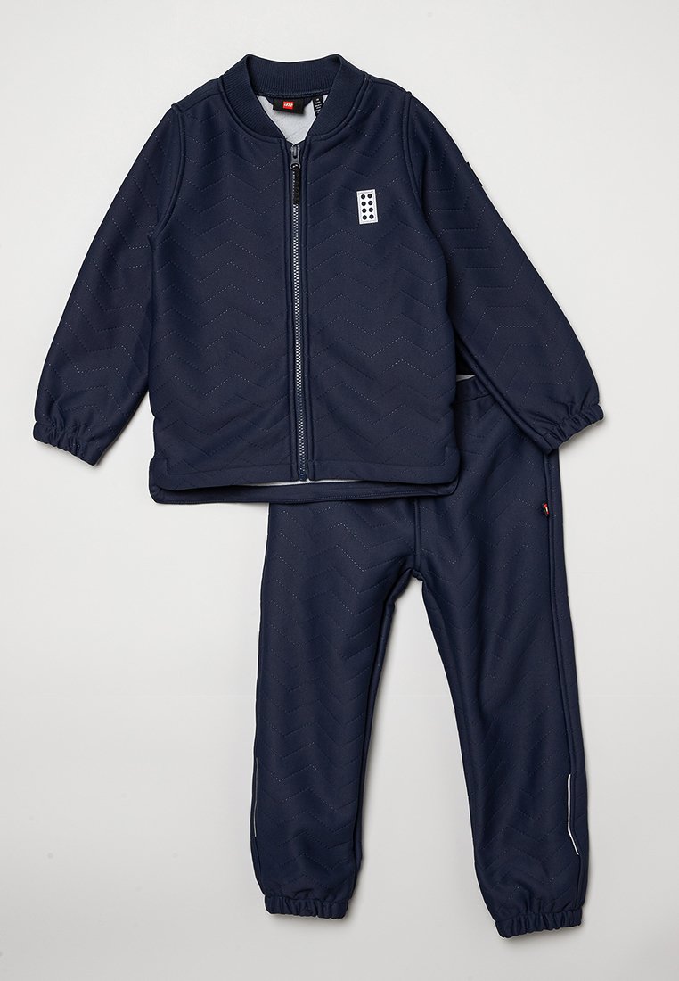 Giacca zip-up blu navy e pantaloni coordinati in tessuto testurizzato, con un motivo a zig zag e polsini elastici, accompagnati da un piccolo logo decorativo.