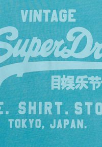 Lichtblauwe shirt met opvallende witte tekst "VINTAGE SuperDry E. SHIRT. STORE TOKYO, JAPAN" in verschillende lettertypes.