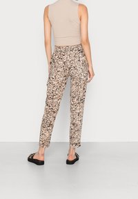 Pantalon imprimé beige avec motifs abstraits noirs. Coupe slim, taille élastique, jambes droites. Porté avec des sandales plates noires.