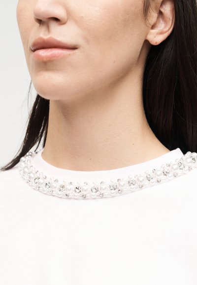 Κοντινό πλάνο ενός ατόμου που φοράει λευκό πουκάμισο με λαιμόκοψη διακοσμημένη με πέρλες και διάφανα στρας.