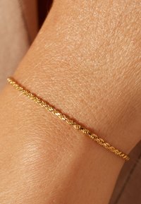 Gouden armband met een gedraaide design. Het ligt op een pols met zichtbare textuur en toont een fijne, delicate uitstraling.