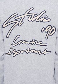 Ανοιχτό γκρι φούτερ με μεγάλα, στυλιζαρισμένα ναυτικά μπλε γράμματα στην πλάτη. Το κείμενο αναγράφει "Stulli '90 Creative Departments" με ρευστό καλλιγραφικό στυλ.
