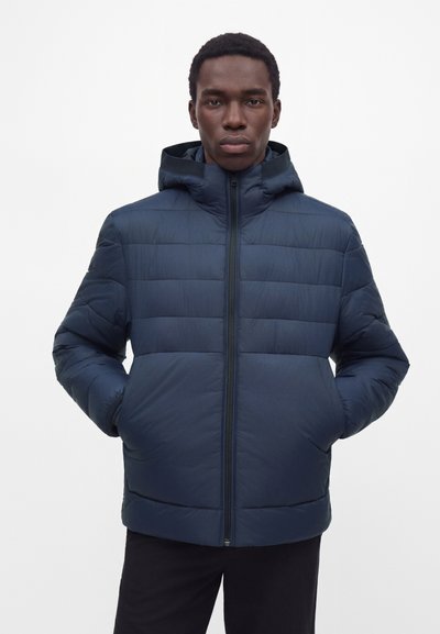 Homme portant une veste matelassée à capuche bleu marine, les mains dans les poches, debout devant un fond blanc uni.