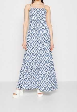 Vrouw draagt een maxi-jurk met een blauw-wit patroon, dunne bandjes en witte platformsandalen, staand tegen een effen achtergrond.