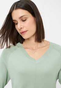 Maglione verde chiaro a scollo a v con una finitura testurizzata e cuciture sulle spalle sottili, abbinato a collane circolari in argento a strati.