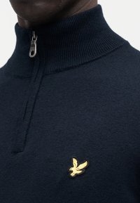 Laivastonsininen neulottu paita suppilokauluksella, jossa on hopeanvärinen vetoketju ja kultakirjailulla koristeltu kotkalogo vasemmassa rinnassa. Pehmeä rakenne.