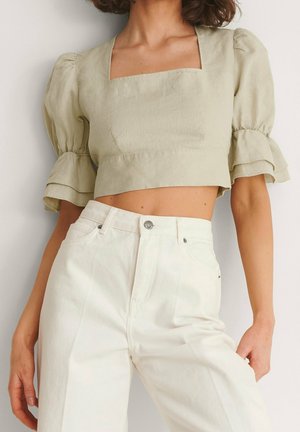Kobieta ubrana w beżowy krótki top z kwadratowym dekoltem, bufkami, warstwowymi rękawami oraz białe spodnie z wysokim stanem na jednolitym tle.