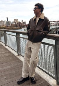 Rjava usnjena bomber jakna s podlogo iz ov�čjega krzna, svetlo teksturirane široke hlače in črni čevlji z debelim podplatom, casualno oblečeni na prostem ob obali.